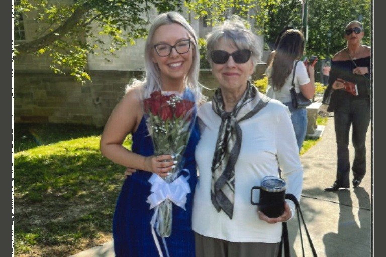 A young woman in blue with flowers stands lovingly next to an older woman with glasses.